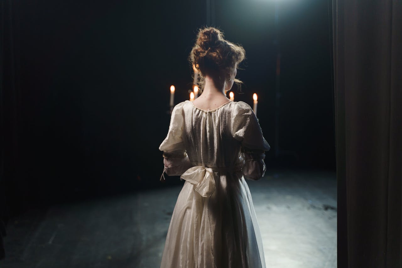 An actress in a white dress stands backstage in dim lighting, enhancing a classic theatrical atmosphere.